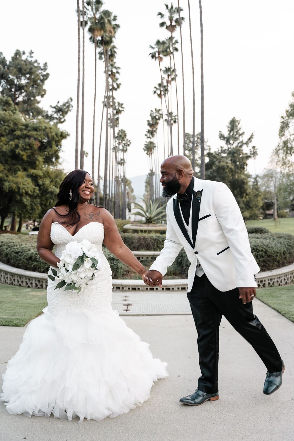 A romantic wedding scene on a palm tree-lined path with the couple holding hands while sharing a joyful moment.