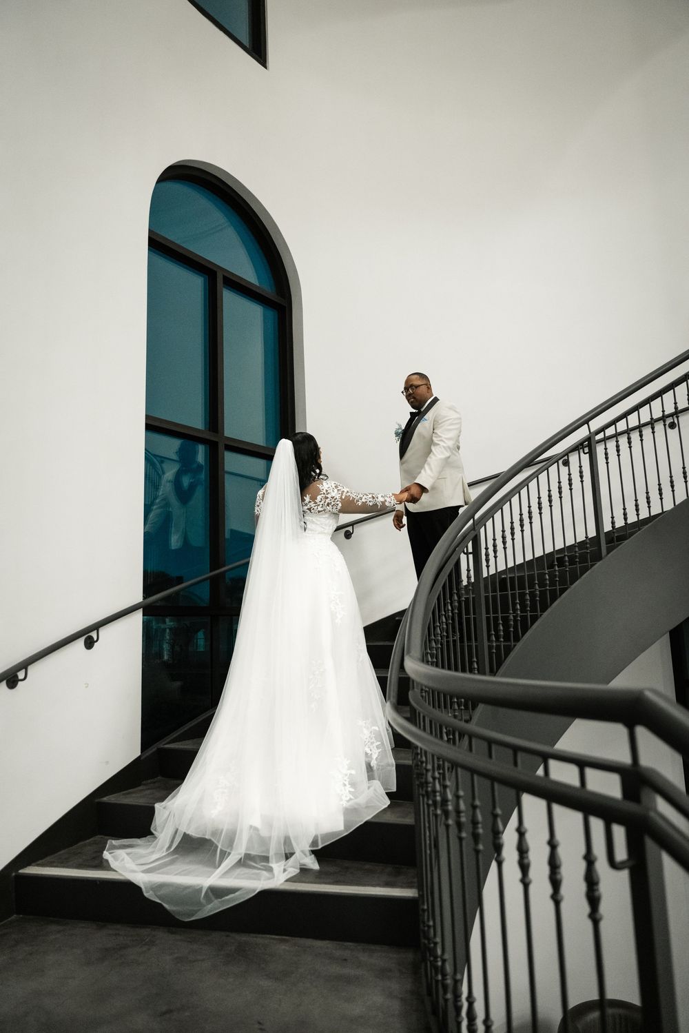 Couple in wedding attire stand on an elegant curved staircase with black railings beside a large arched window.