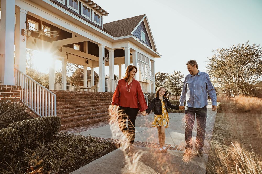 A family walks along a sunset-lit path in front of a charming craftsman style house with white columns and wraparound porch.