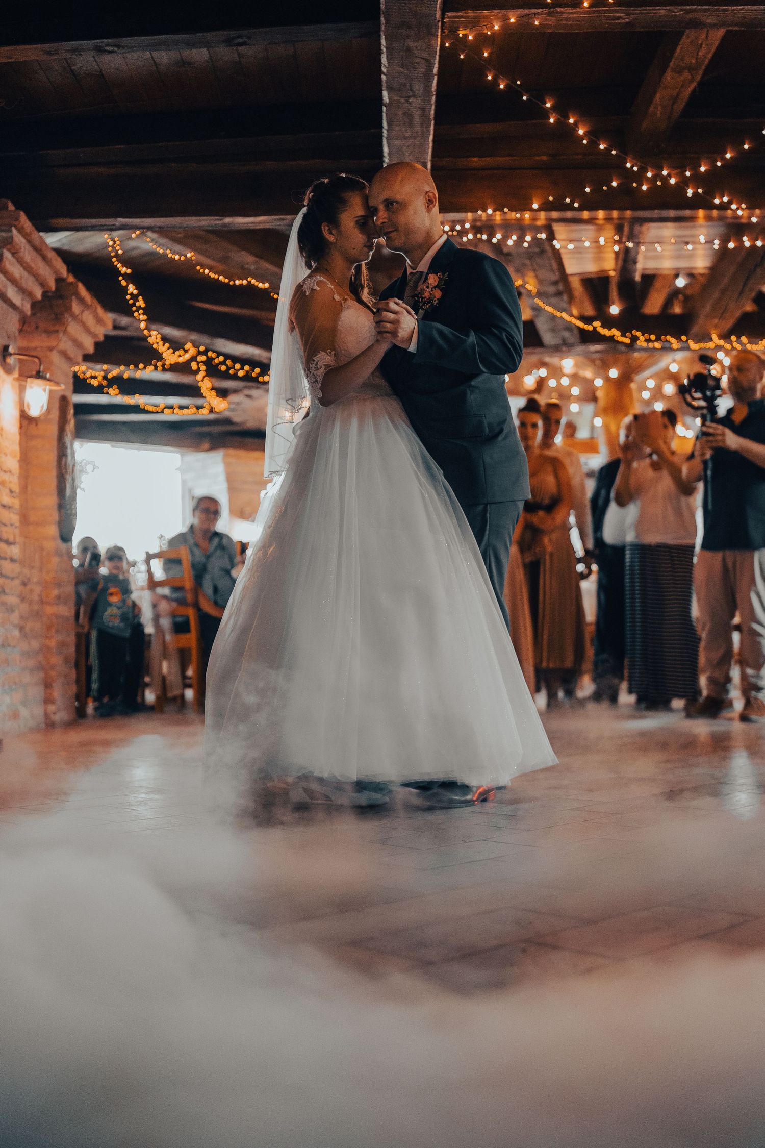 Couple shares their first dance at rustic wedding reception with string lights and romantic fog on dance floor.