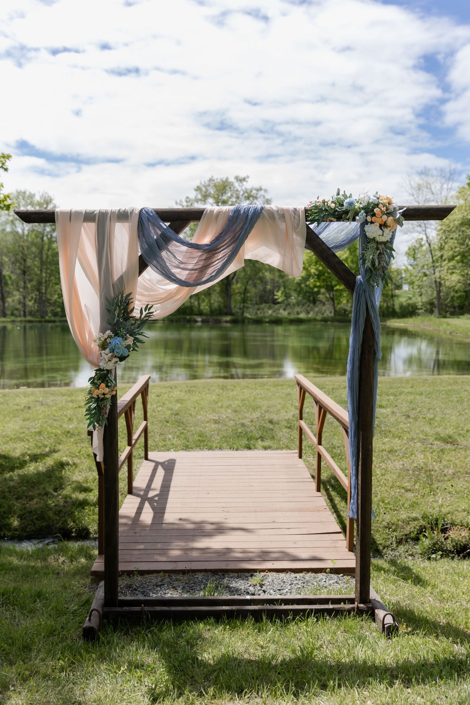 A rustic wooden wedding arch with draped fabric and floral decorations stands on a dock overlooking a serene lake.