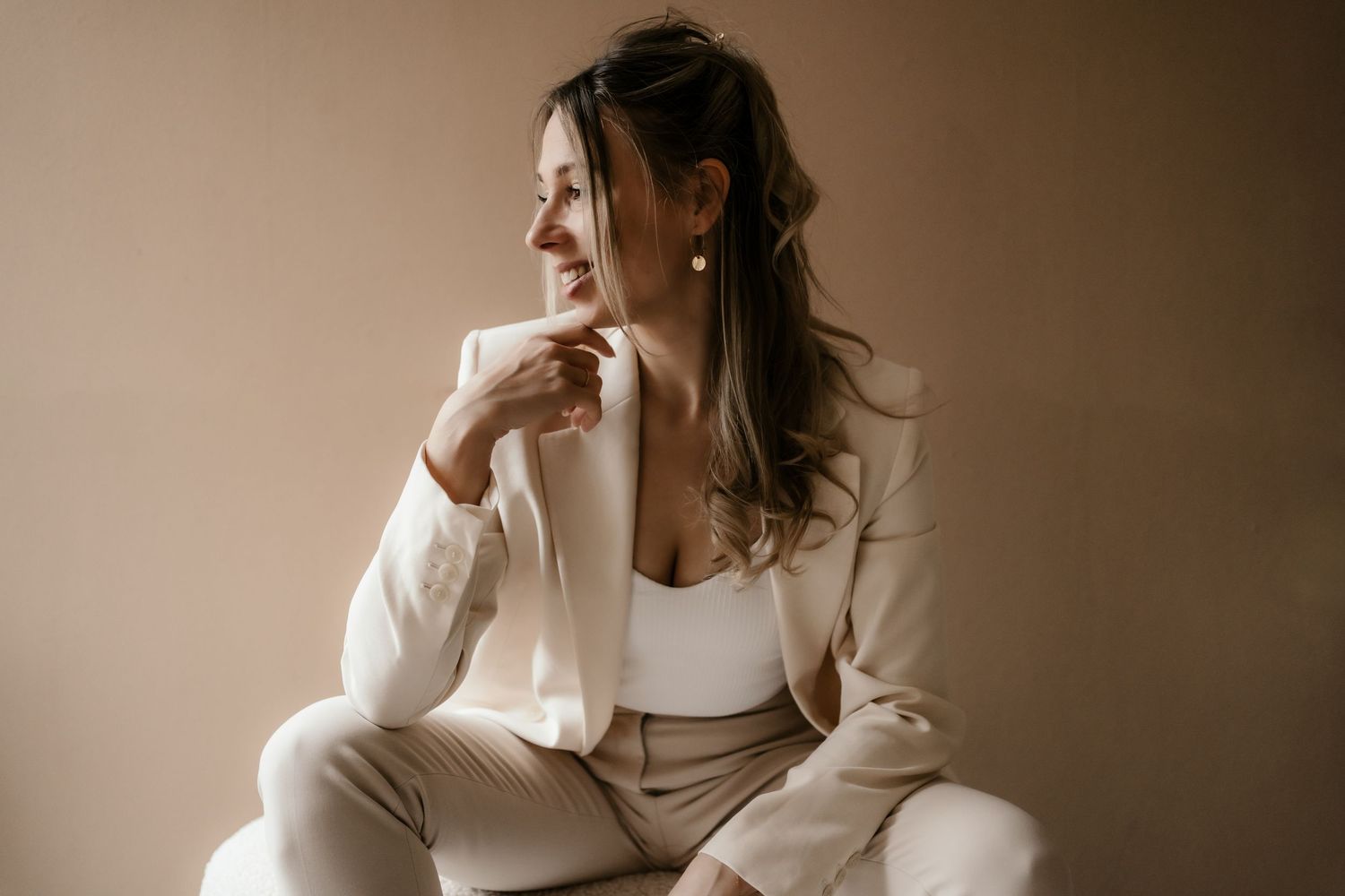 Elegant figure in cream-colored business suit sits thoughtfully against neutral background in soft lighting.