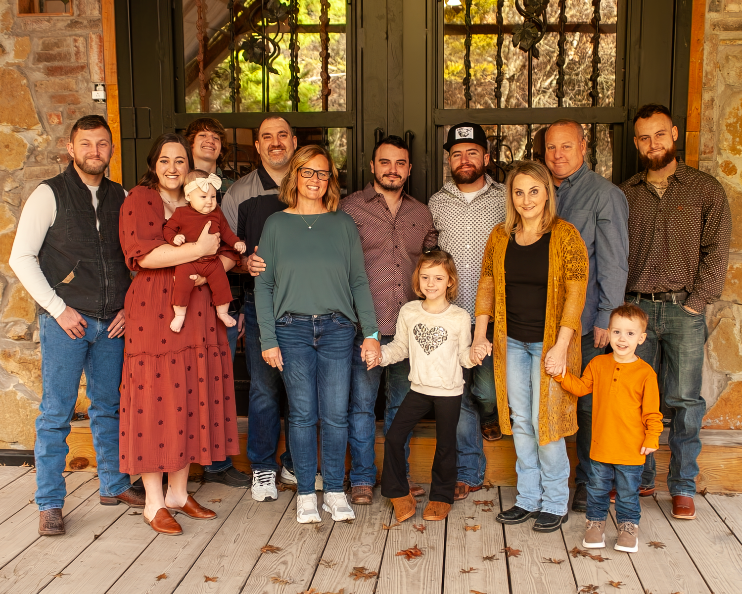 Family group wearing coordinated casual outfits standing together on rustic wooden porch of stone building.