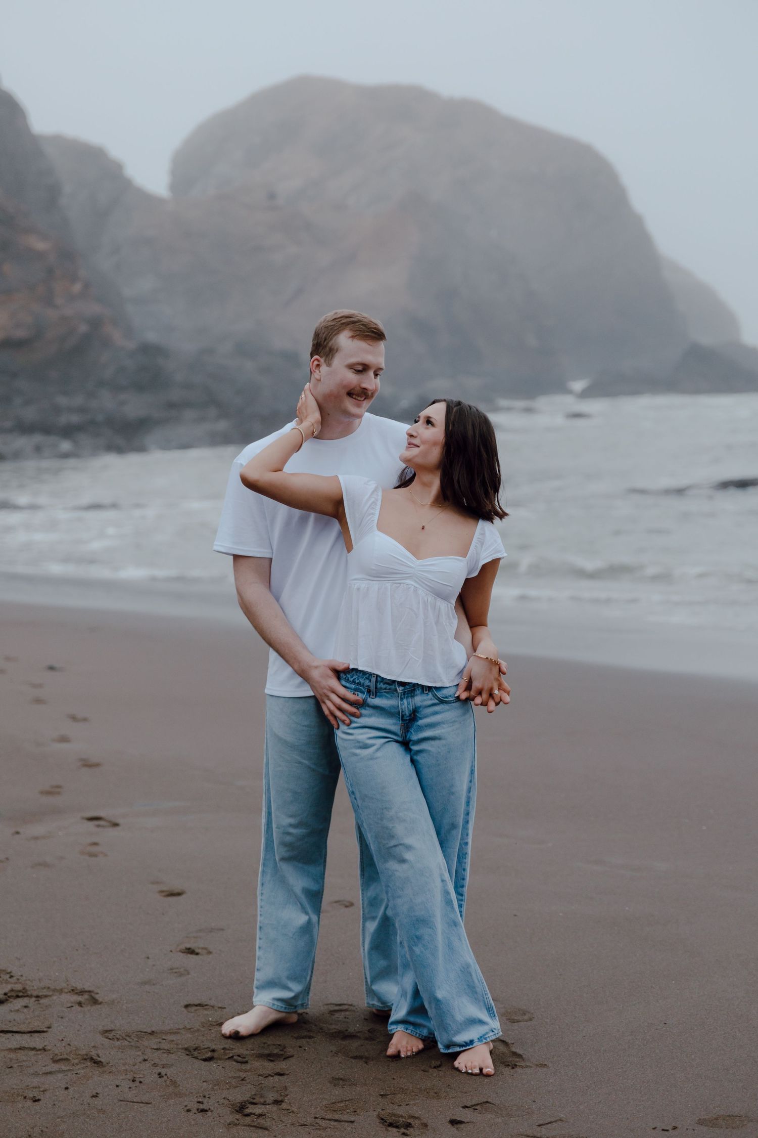 A couple in casual attire embraces on a misty beach with rocky cliffs in the background.