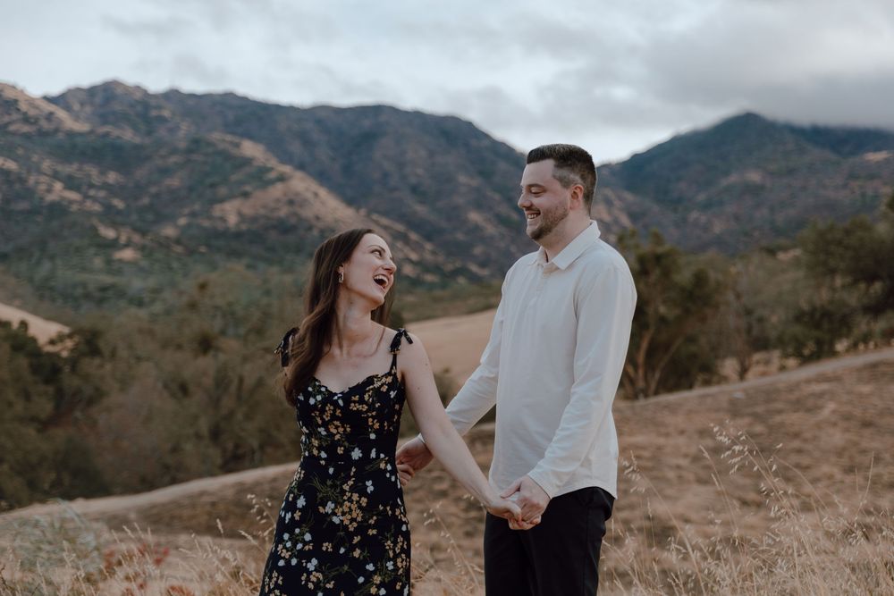A couple holding hands and laughing together on a mountain trail with rugged peaks and cloudy skies in the background.