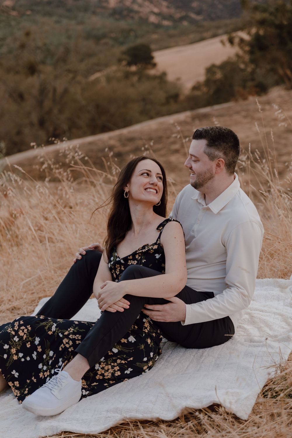 A couple sits together on a blanket during sunset on a hillside, sharing an intimate moment in a natural setting.
