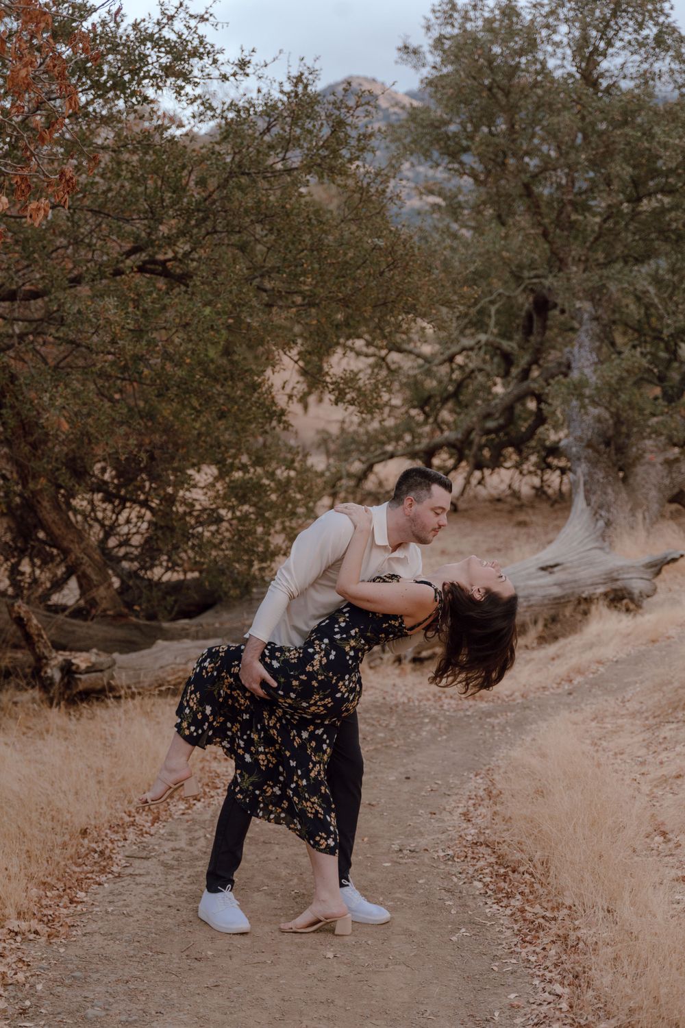 A couple shares a romantic dip pose on a dirt path surrounded by trees during an autumn photoshoot.