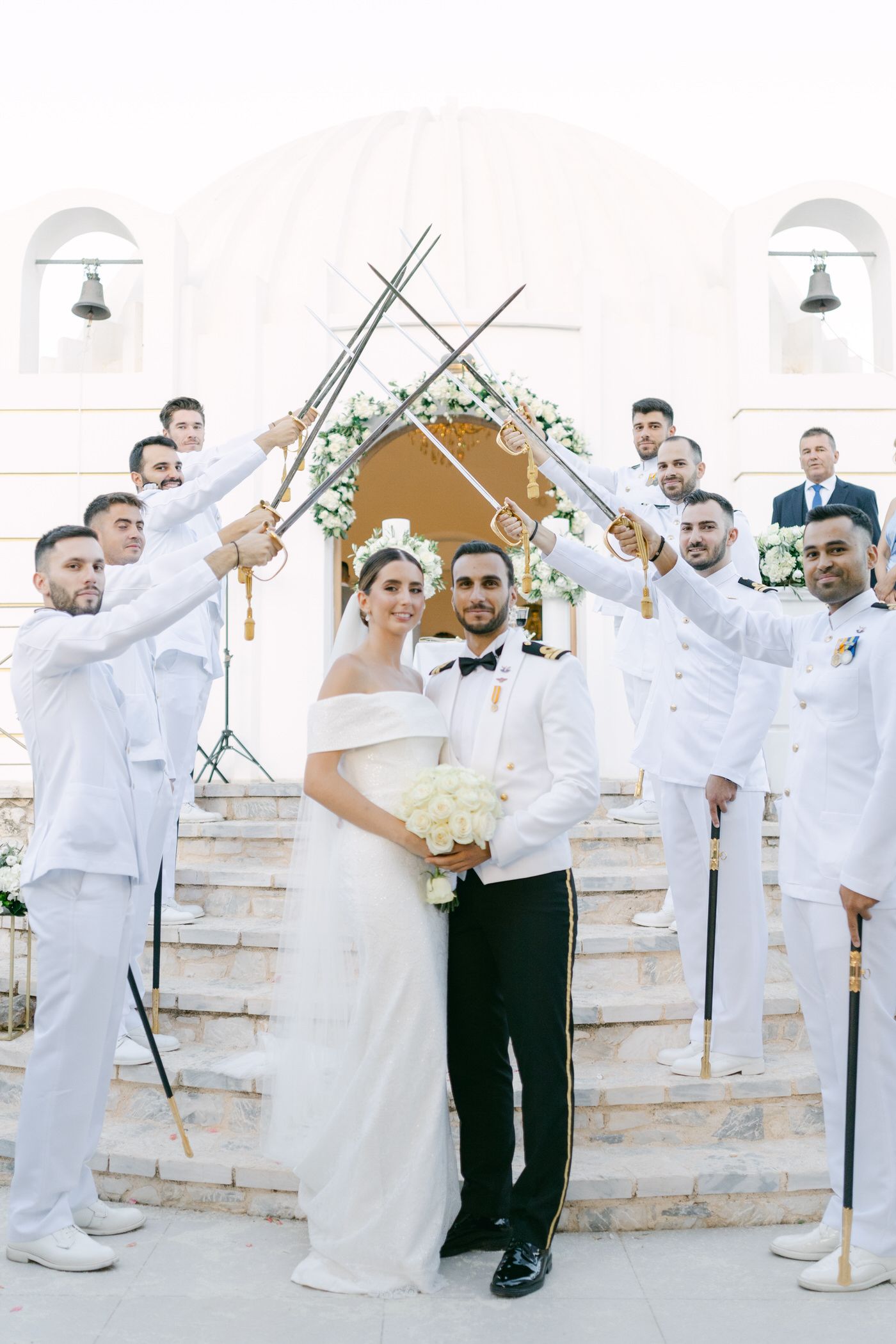 Wedding party in white attire creates an arch of gold swords over couple during outdoor ceremony on stone steps.