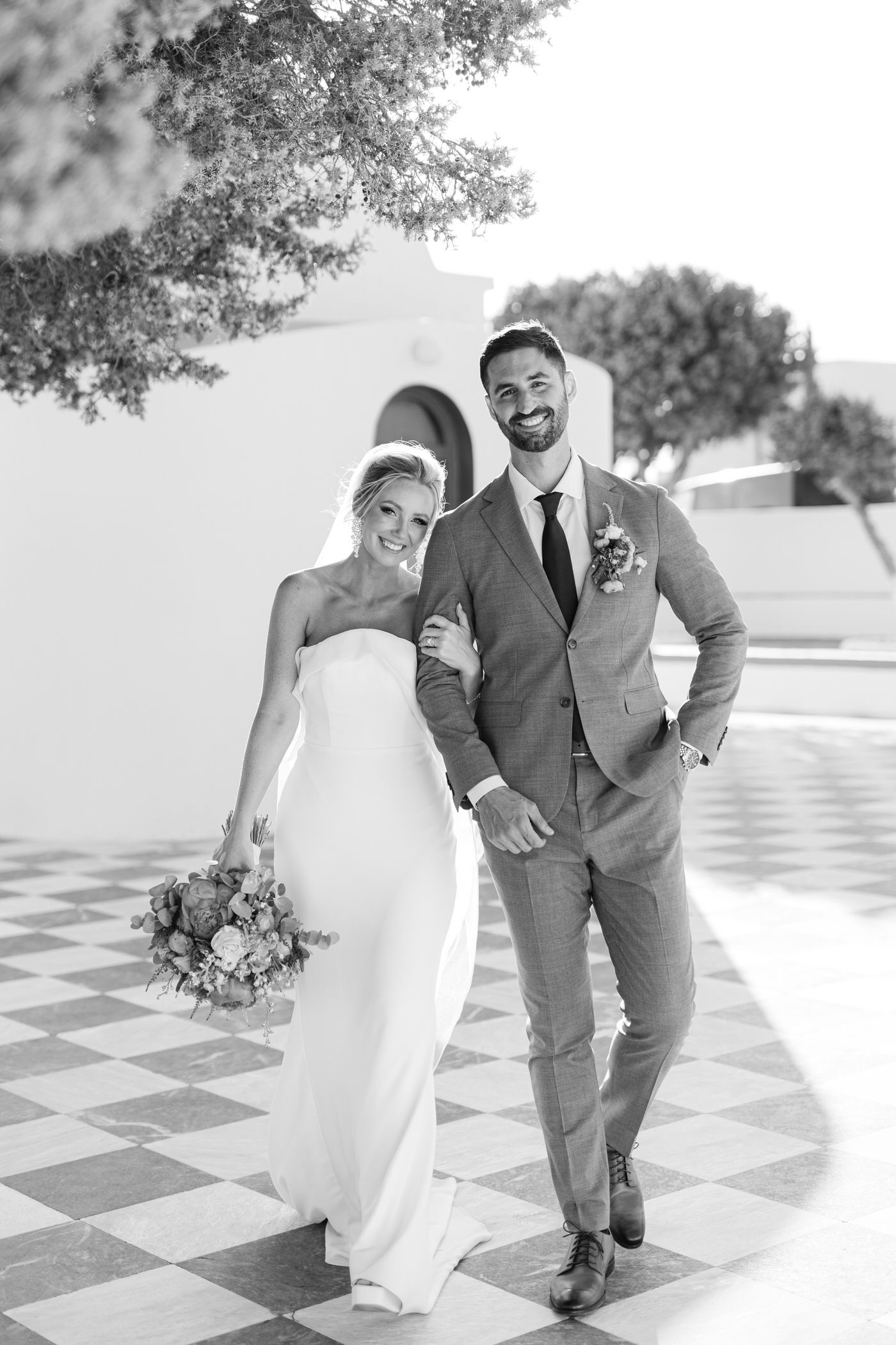 A newlywed couple in formal wedding attire smiles while walking together under dappled sunlight on a checkered patio.