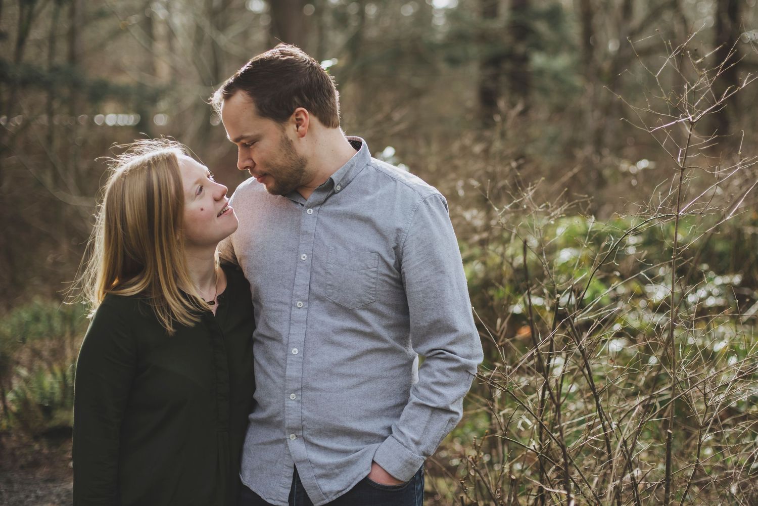 A couple stands close together on a quiet path in the woods, sharing an intimate moment as they gaze into their eyes.