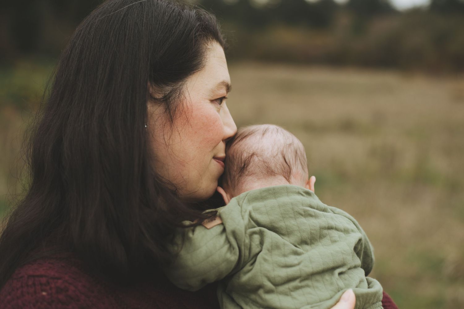 A tender moment captured in profile as a mother gently kisses her baby wearing a green outfit in an outdoor setting.