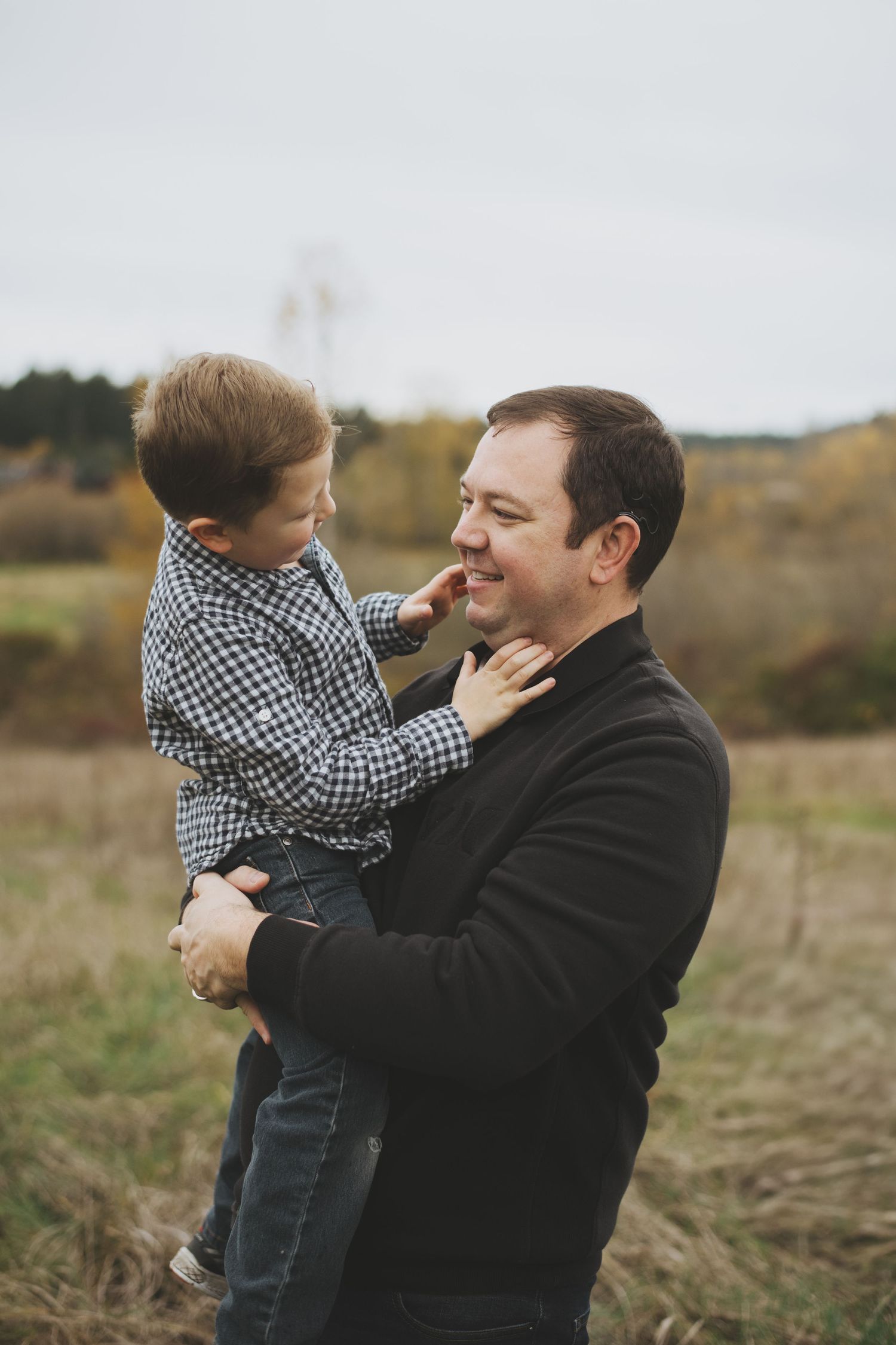 A tender moment between a young boy in plaid and his father in black outdoors in an autumn field.