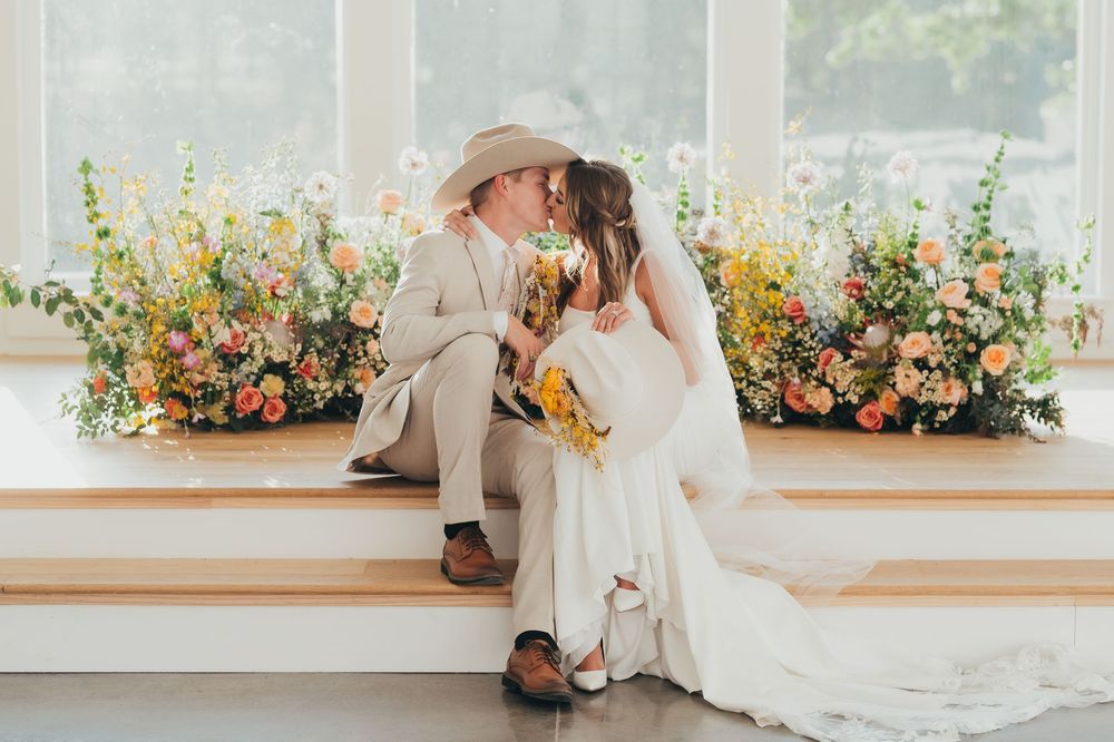 A couple in wedding attire share an intimate moment on steps surrounded by colorful floral arrangements.