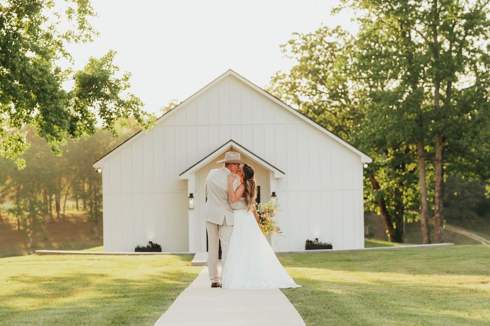 A couple shares an intimate moment outside a white rustic chapel surrounded by green trees in the golden sunlight.