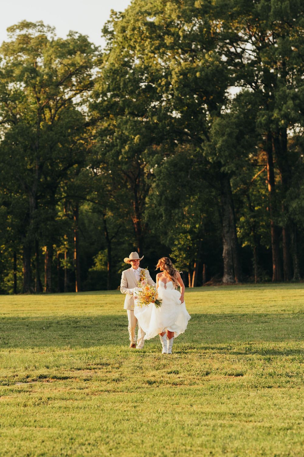 A couple running through a sunlit meadow surrounded by tall trees during their wedding day.