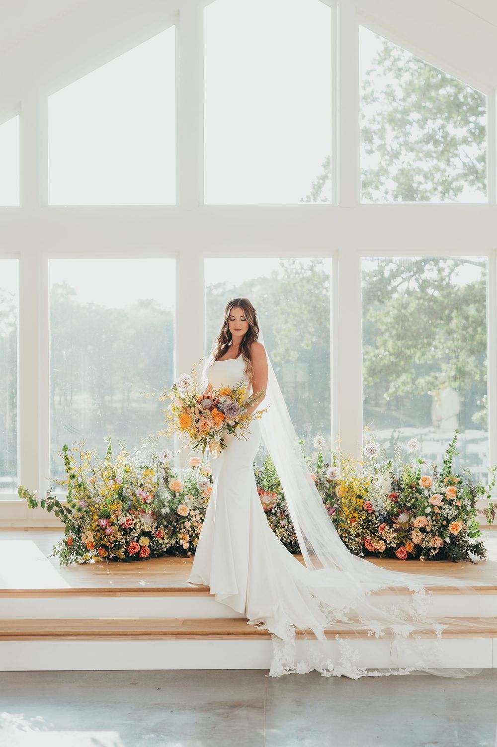 A bride poses in a flowing white gown against large windows with floral arrangements.