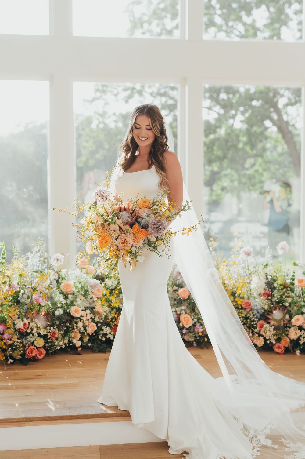 Elegant bride in sleek white mermaid gown holds vibrant peach and orange bouquet against floral wall backdrop in sunlit venue.