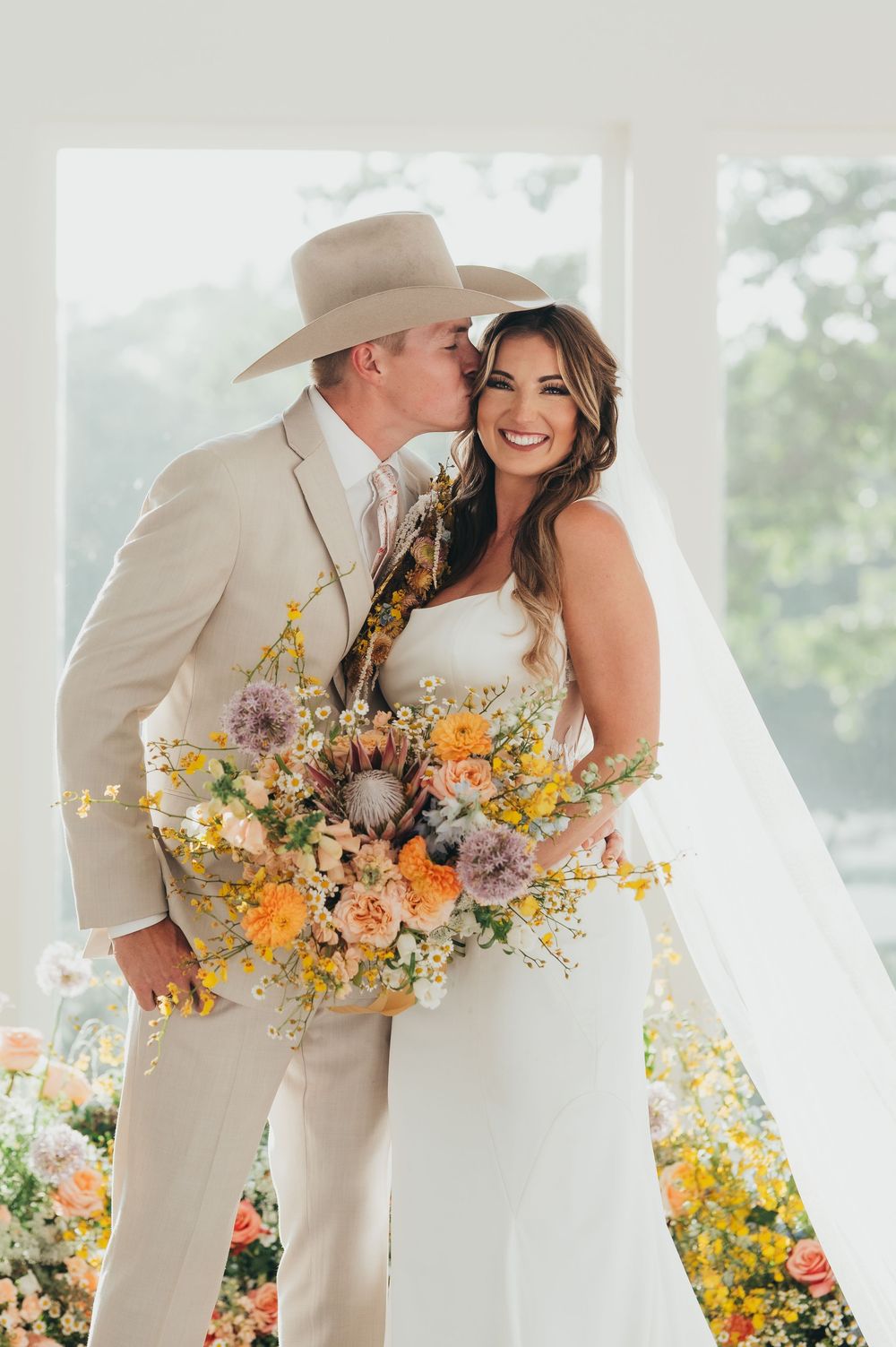 A wedding couple embraces in tan and white attire with a vibrant wildflower bouquet against a bright outdoor setting.