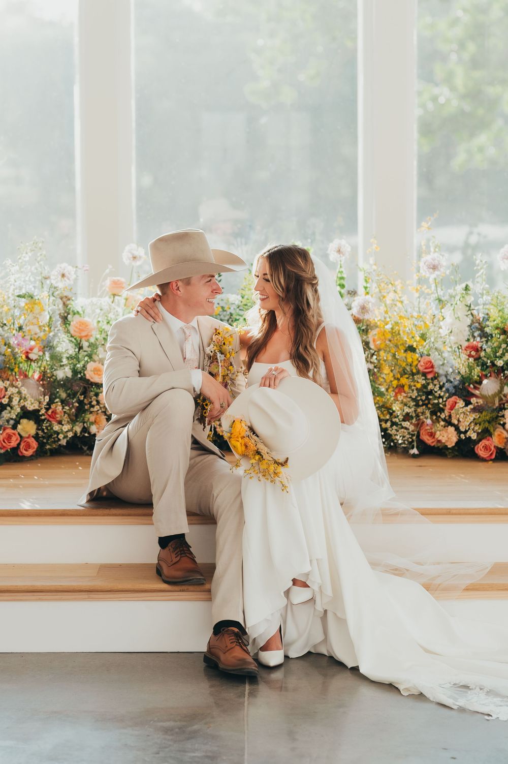 A wedding couple sits intimately on steps with a beautiful floral backdrop during their rustic-themed celebration.