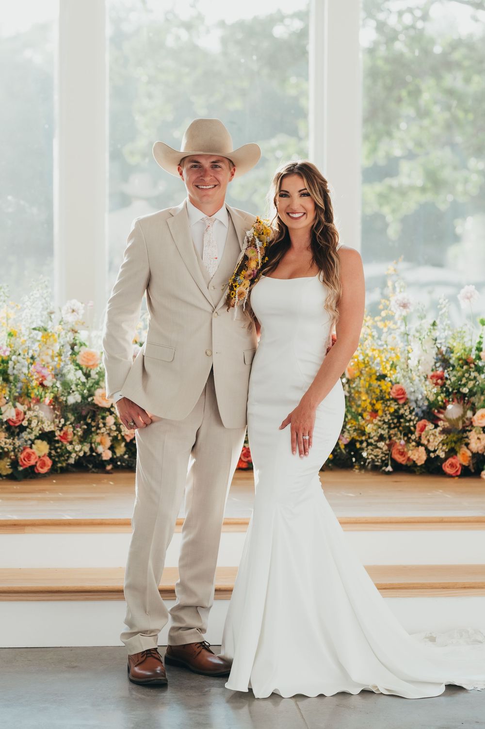 A wedding couple poses in front of a colorful flower wall, wearing a beige suit and white mermaid dress.