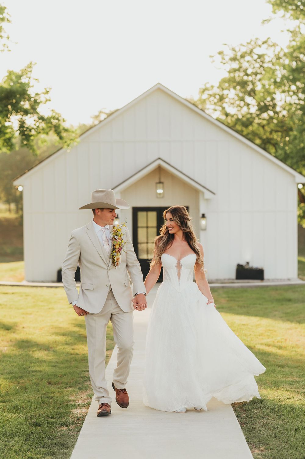 A romantic wedding couple walking together on a path in front of a white chapel during golden hour.
