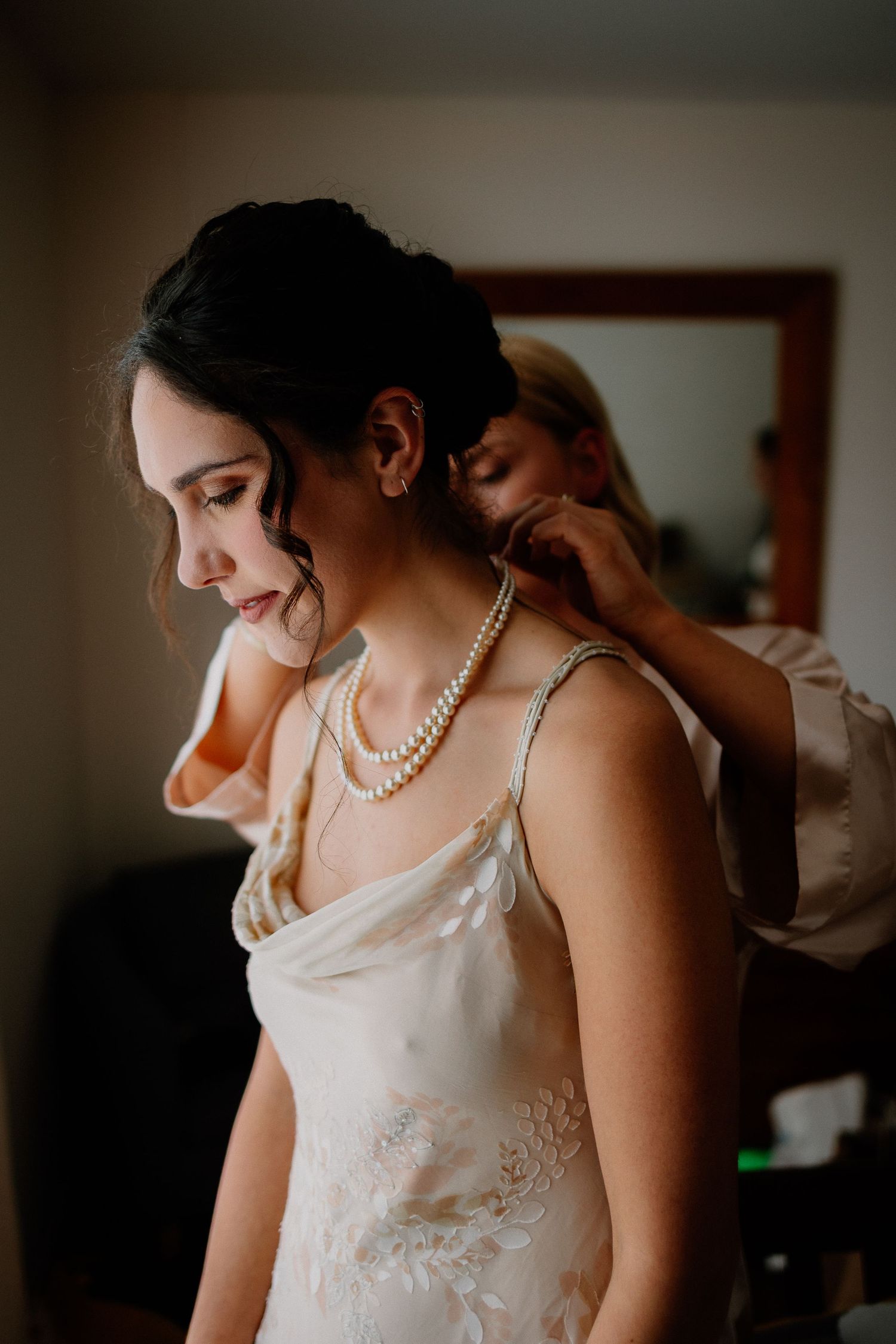 Someone wearing a pearl necklace and white lace dress smiles while getting ready in soft moody lighting.