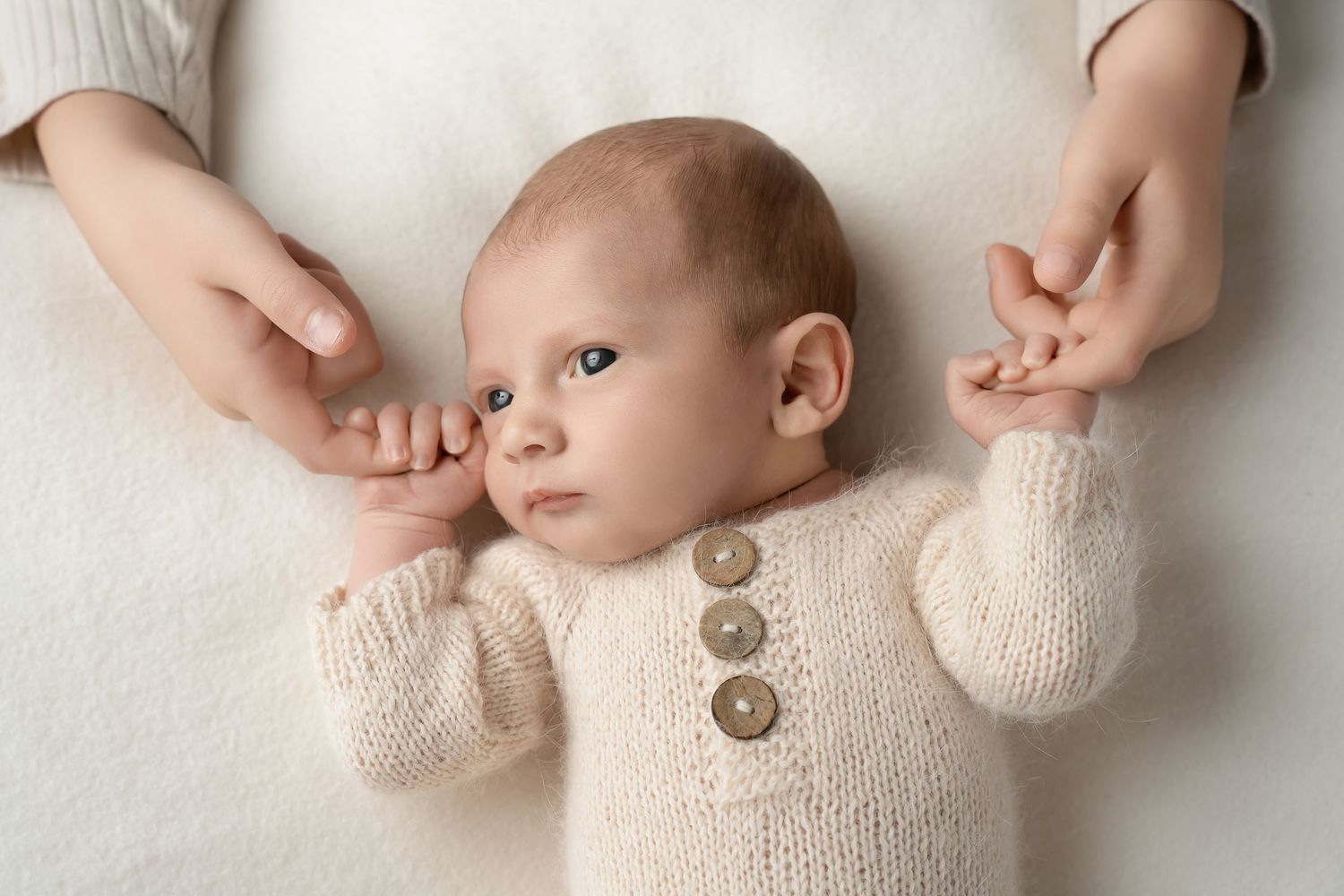 Baby wearing cream knit sweater with wooden buttons lies on white blanket while hands gently hold their arms.