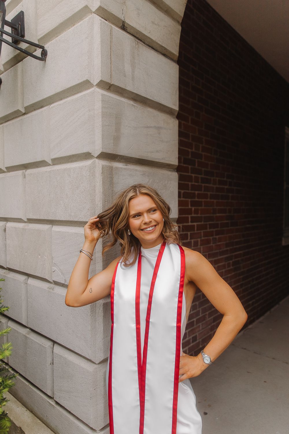 A graduate in a white dress with red graduation stoles poses against a stone and brick building exterior.