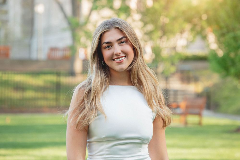 A person in a white sleeveless dress smiles while standing outdoors in a park-like setting on a sunny day.
