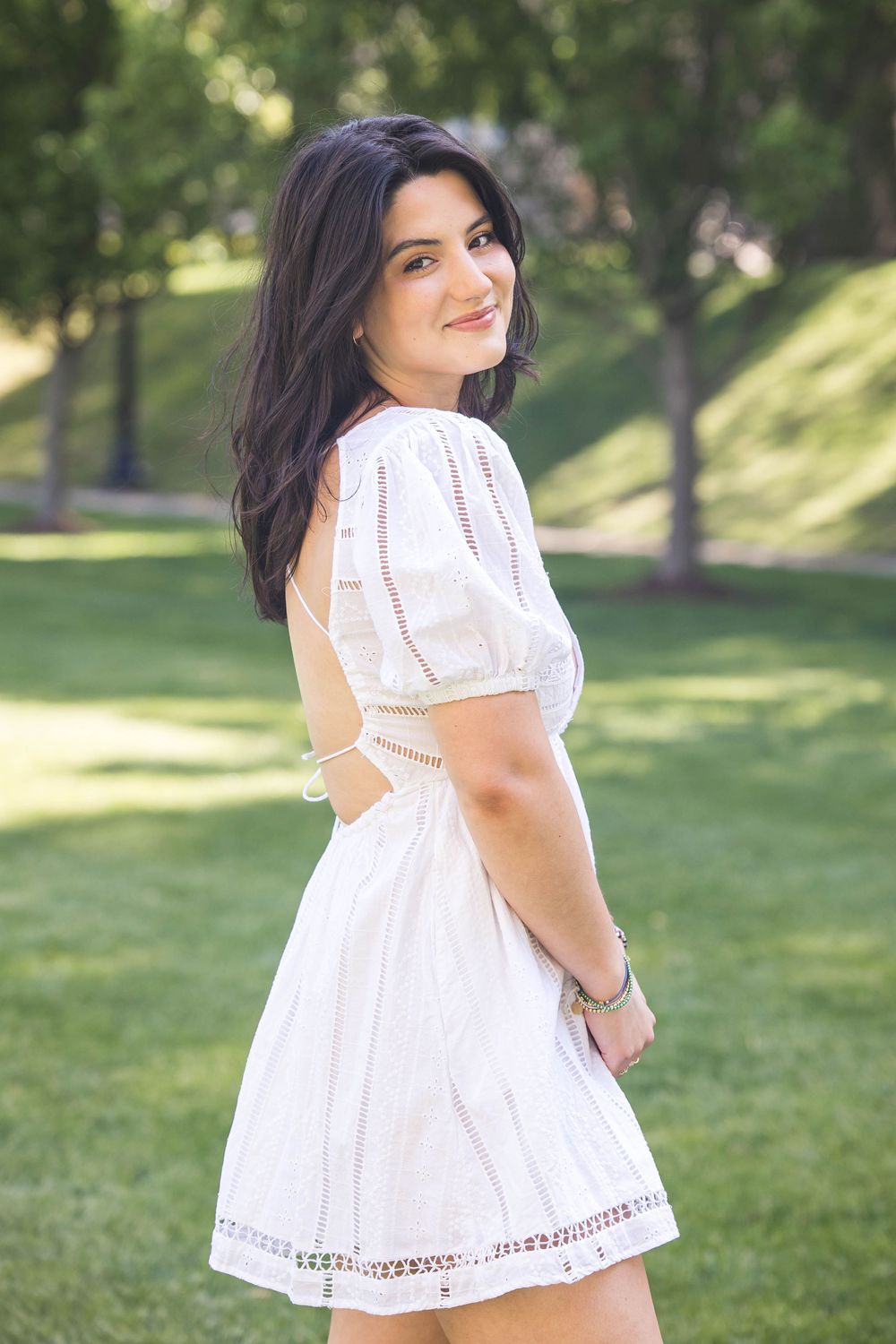 A person in a white summer dress poses outdoors in a sunny park setting with lush green grass and trees.