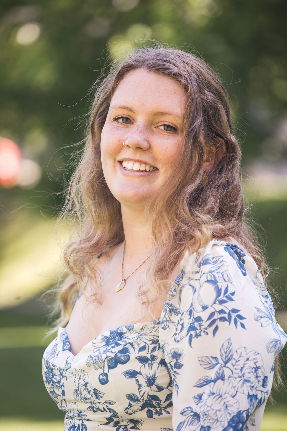 A person wearing a blue and white floral dress smiles while standing outdoors with a blurred natural background.