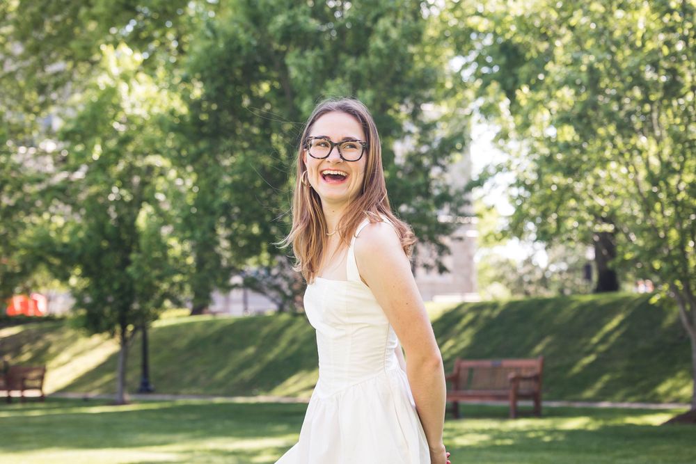 A person in a white sundress and glasses stands smiling in a sunny park with green trees and benches in the background.