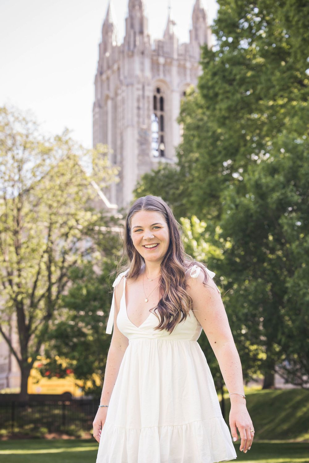 A person wearing a white dress stands smiling in front of a Gothic cathedral tower with trees in the background.