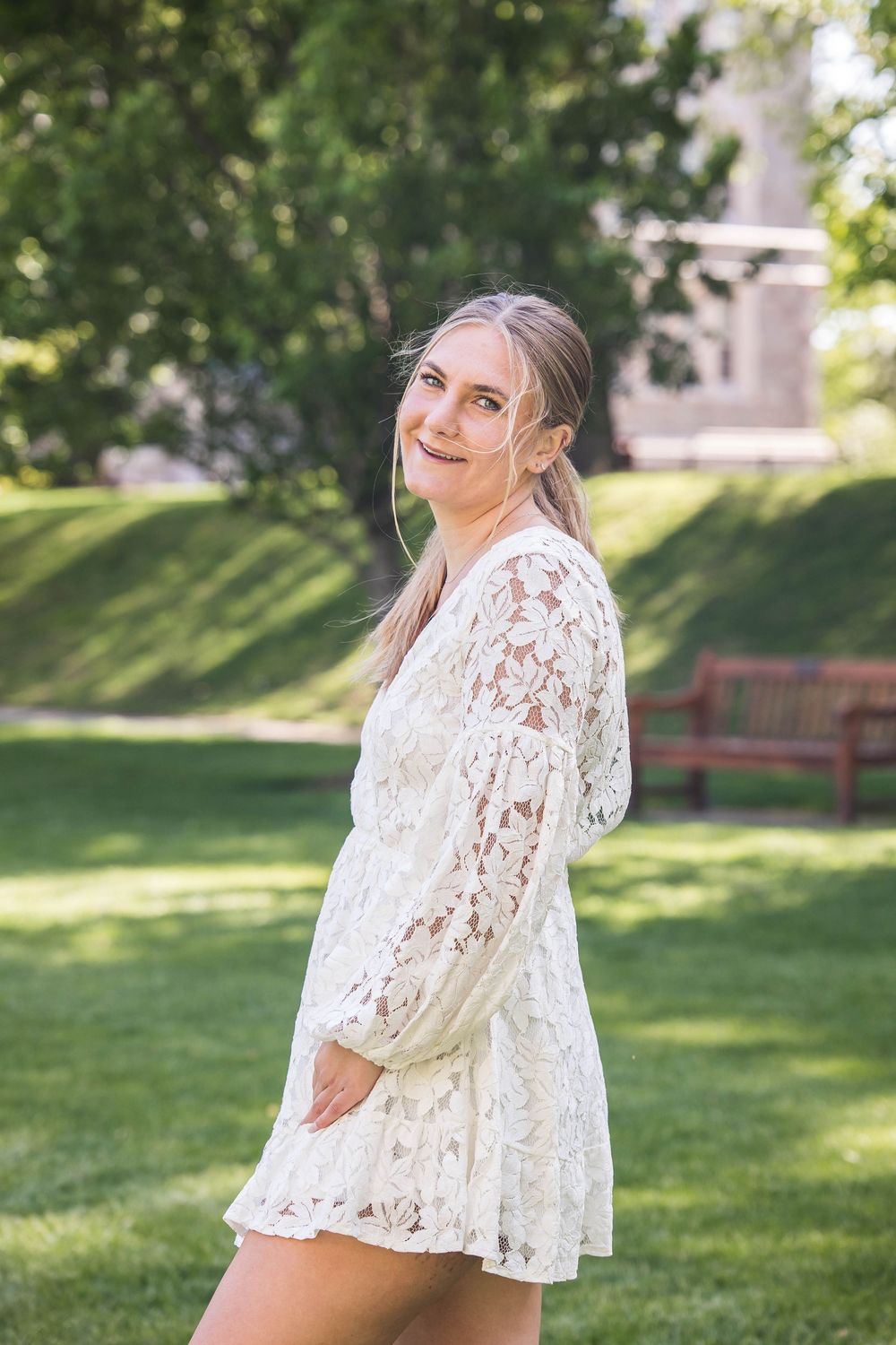Young woman in white lace dress poses outdoors in a sunny garden setting with green grass and trees.