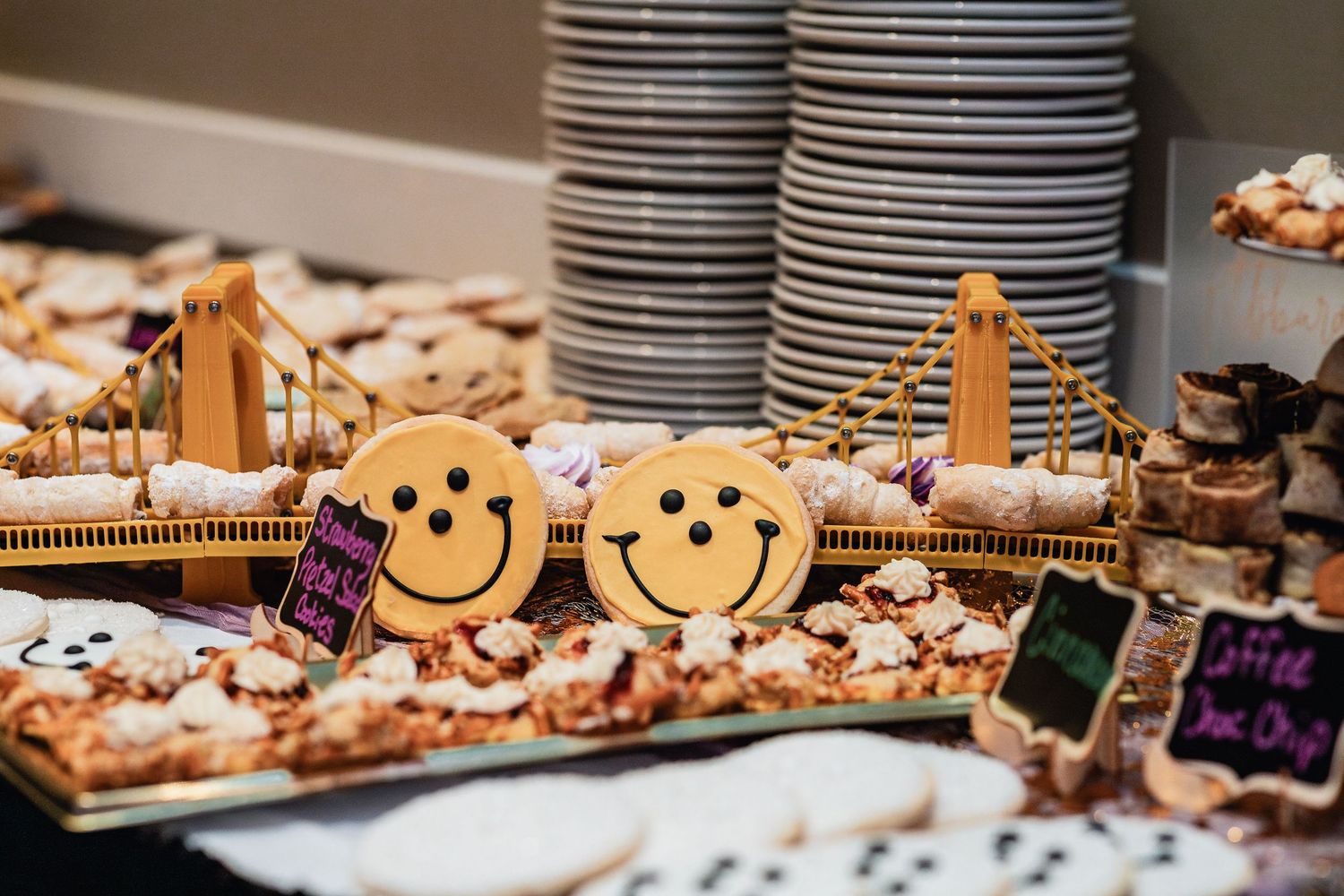 Smiling cookie decorations on display at a catering event with stacked plates and desserts in the background.