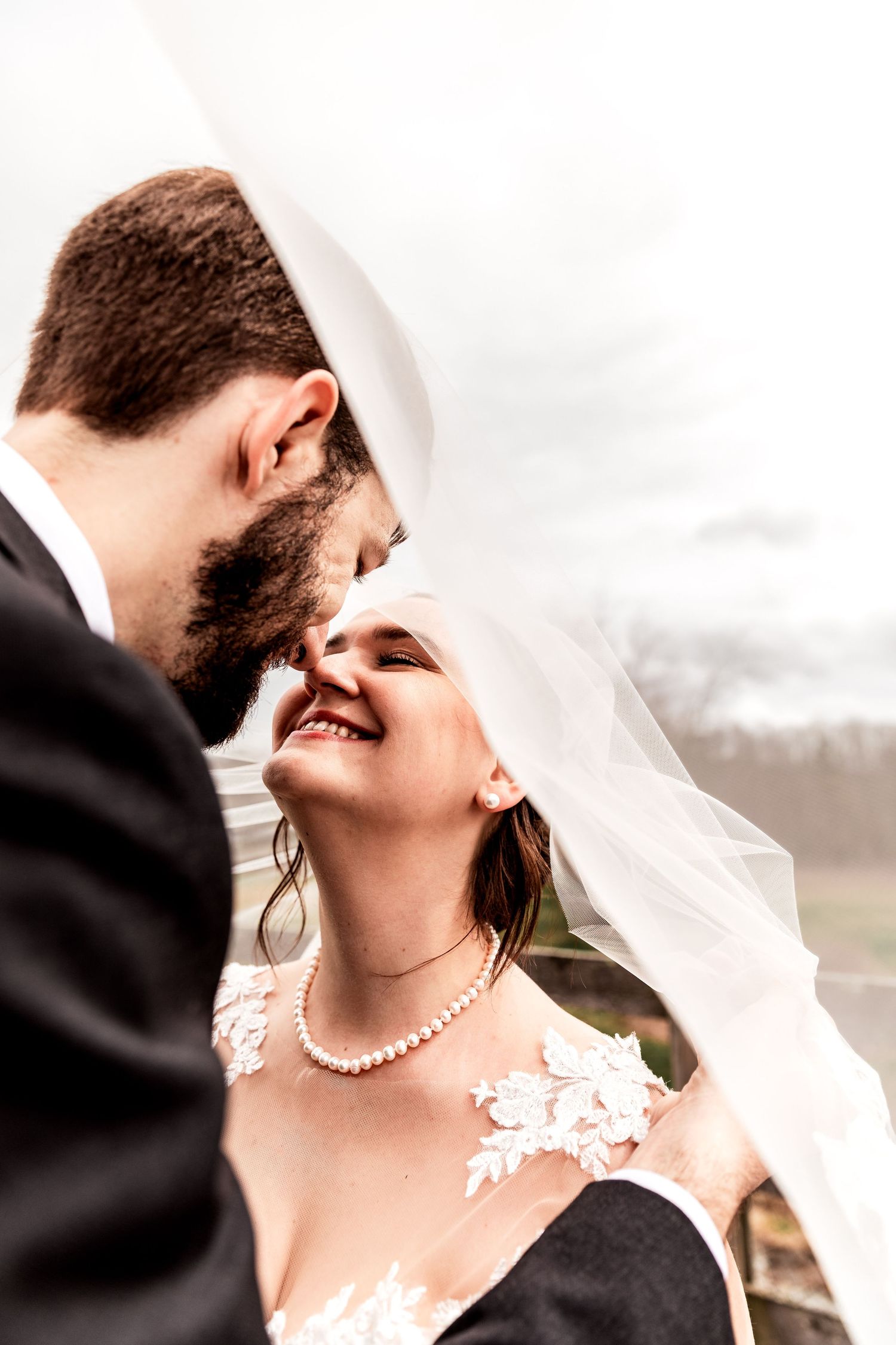 Romantic wedding portrait of bride and groom under veil, sharing an intimate moment outdoors, smiling with joy and love.
