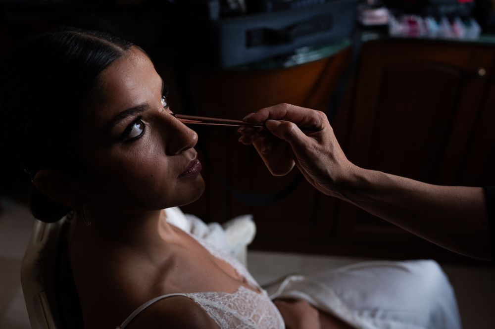 Atmospheric lighting captures the application of eye makeup during wedding preparation.