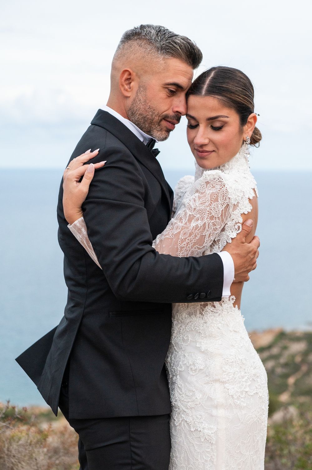 Elegant couple in wedding attire embrace against coastal mountain scenery.