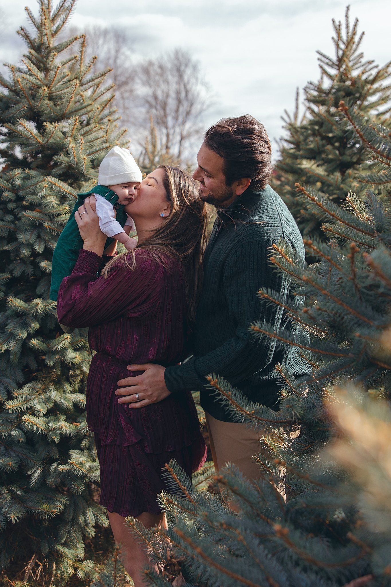 Family shares tender moment among evergreen trees at Christmas tree farm on cloudy winter day.