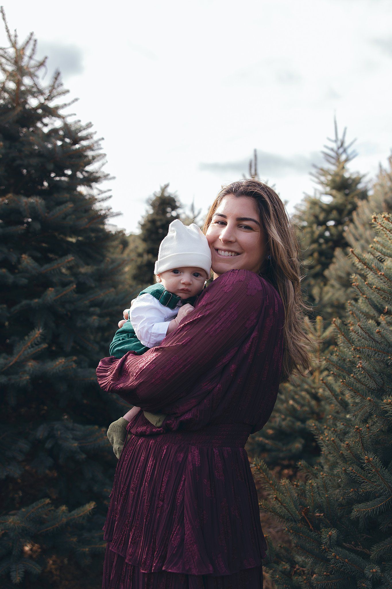 Mother in burgundy dress holds baby in white hat among pine trees on winter day.