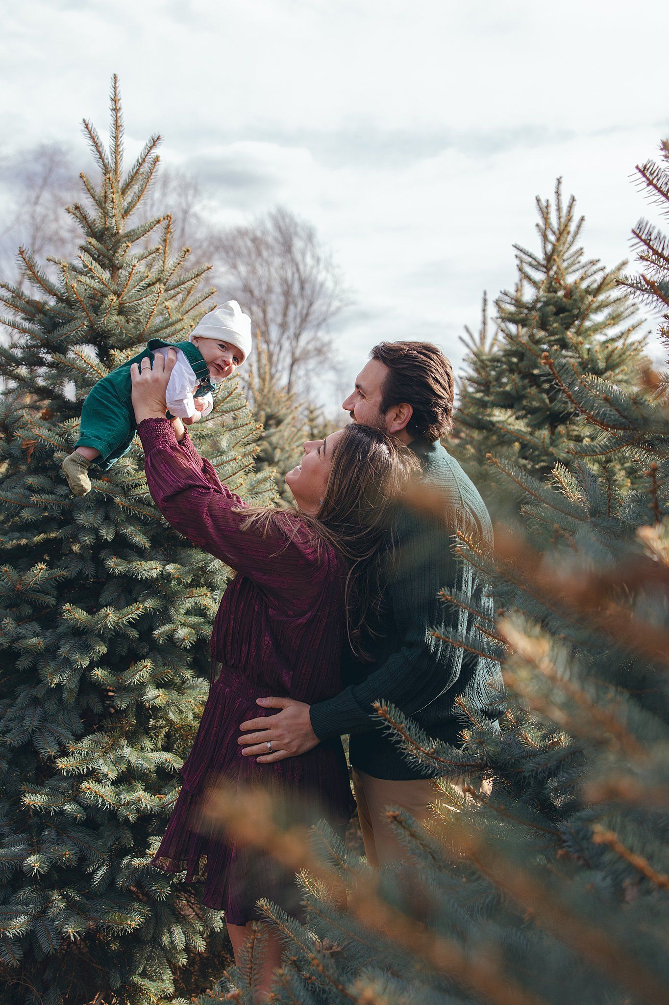 Parents joyfully lift infant in the air surrounded by evergreen trees at tree farm.