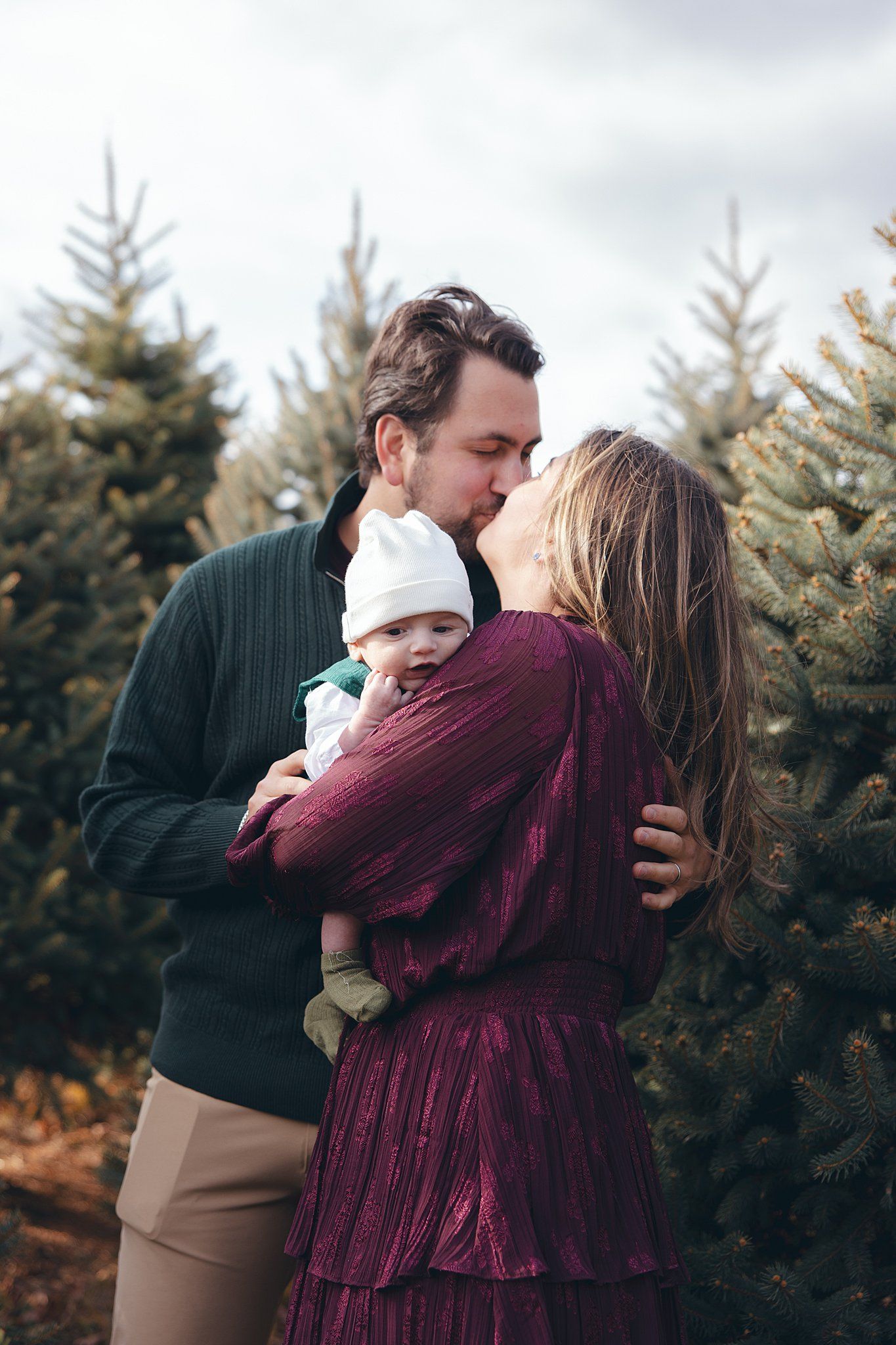 Intimate family moment captured between parents and baby surrounded by Christmas trees.