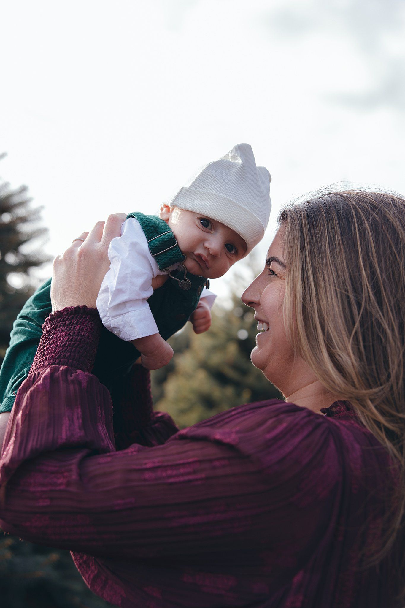 Mother in burgundy dress playfully lifts smiling baby wearing white hat outdoors.
