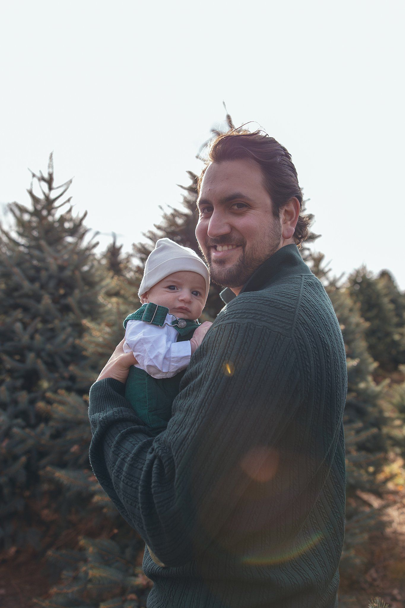 A person holds a small baby while smiling among evergreen trees on a cloudy day.