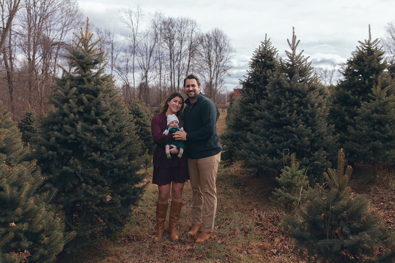 Family of three poses together between rows of evergreen trees at tree farm.