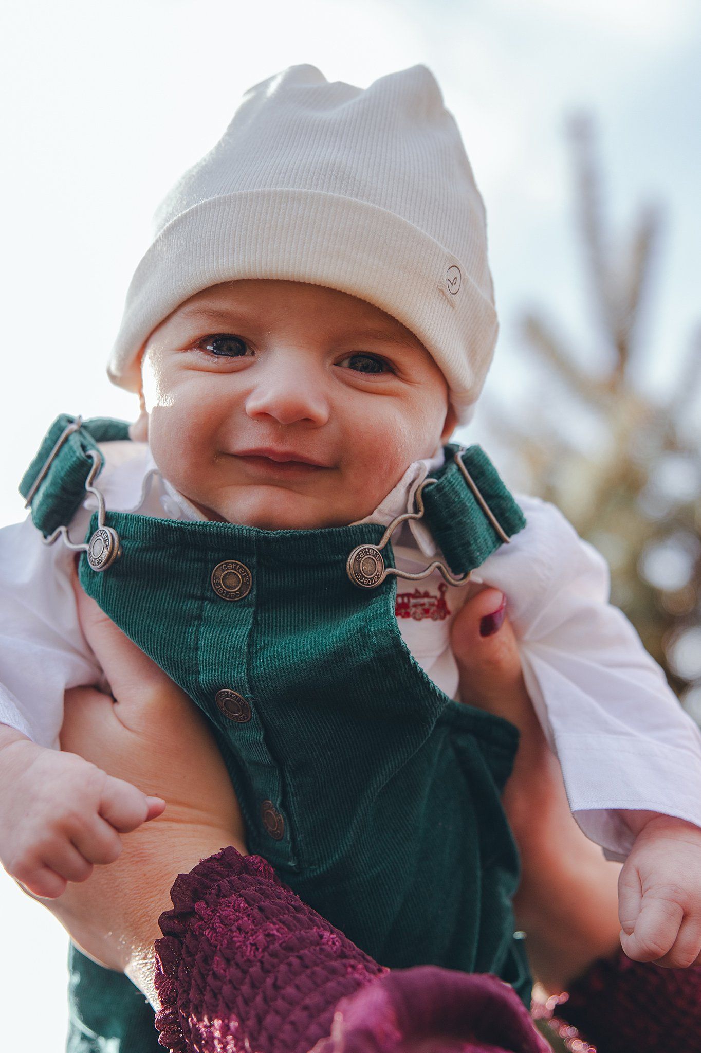 Smiling baby wearing white hat and green overalls in close-up portrait.