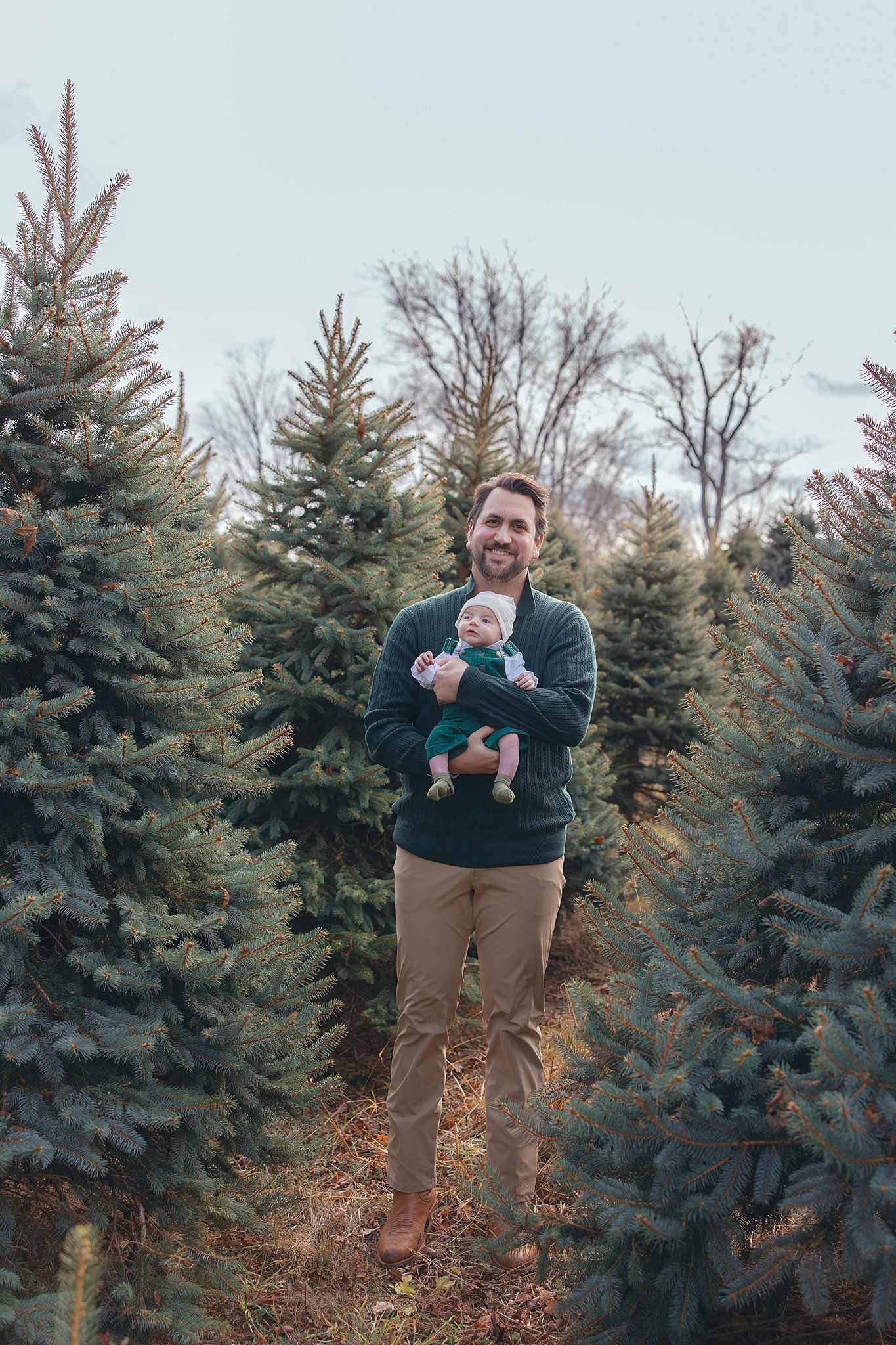 Person stands among evergreen trees holding small baby on winter day.