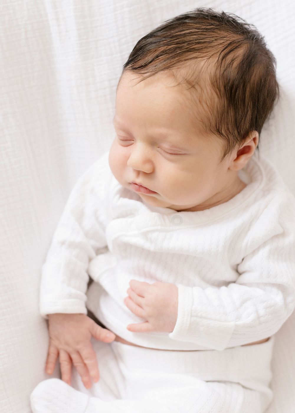A sleeping newborn baby wearing a white outfit rests peacefully against a light-colored background.
