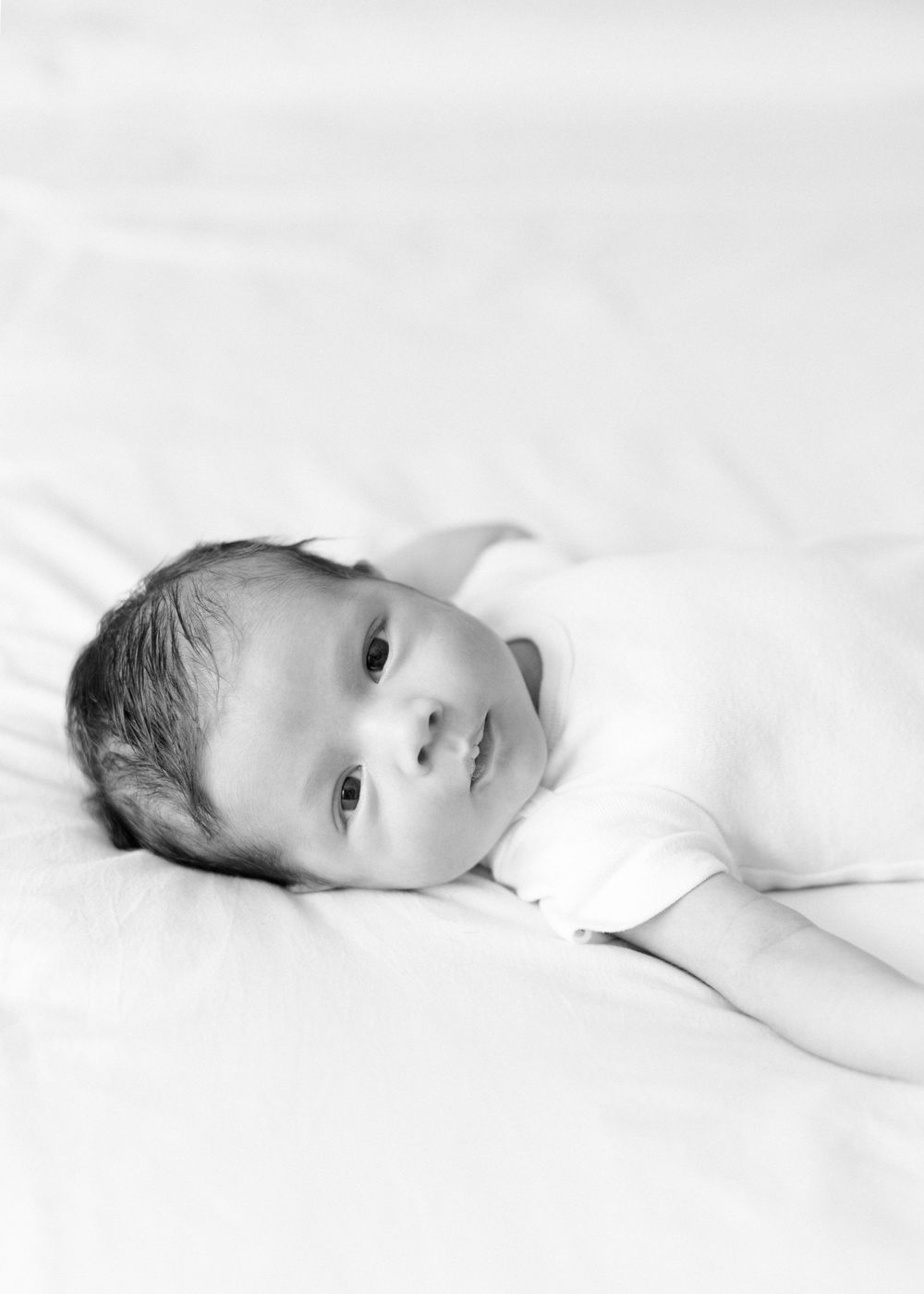 Black and white portrait of a baby lying on white bedding with a watchful expression.