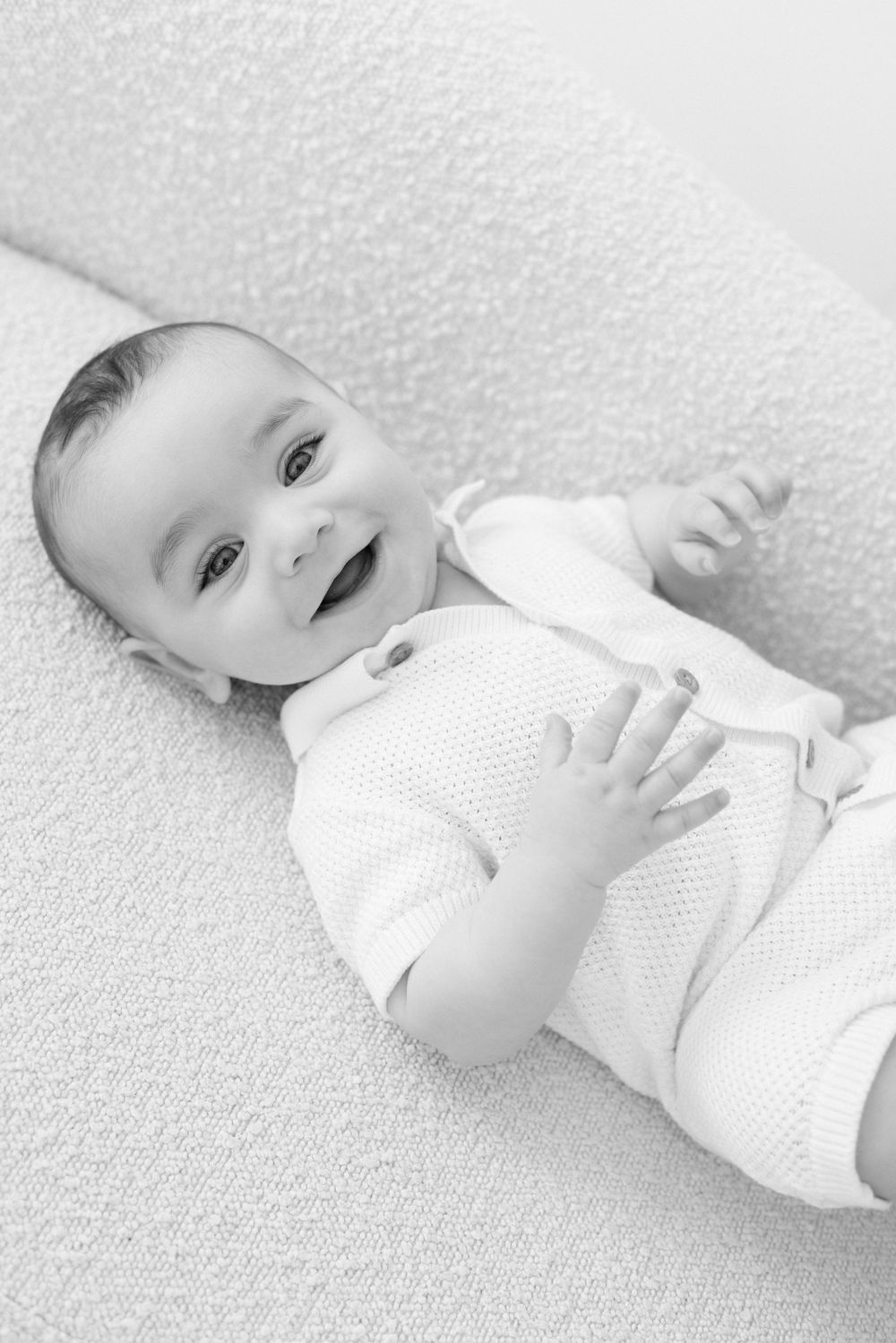 Black and white portrait of a smiling baby lying on textured fabric wearing a white knit outfit.
